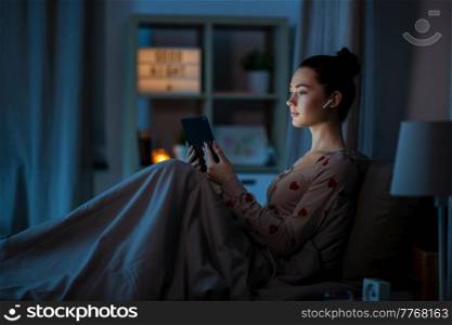 technology, bedtime and people concept - teenage girl with tablet pc computer and earphones sitting in bed at home at night. teenage girl with tablet pc and earphones in bed