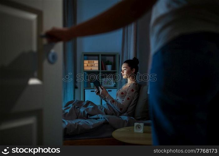 technology, bedtime and people concept - teenage girl with tablet pc computer sitting in bed at home at night and mother entering room. teenage girl with tablet pc in bed at night