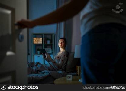 technology, bedtime and people concept - teenage girl with tablet pc computer sitting in bed at home at night and mother entering room. teenage girl with tablet pc in bed at night