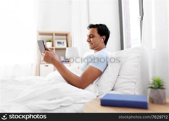 technology, bedtime and rest concept - happy smiling indian man in earphones with tablet pc computer lying in bed and listening to music at home. happy indian man with tablet pc in bed at home