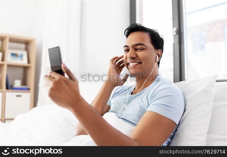 technology, bedtime and rest concept - happy smiling indian man in earphones with smartphone lying in bed and listening to music at home. indian man in earphones with phone in bed at home