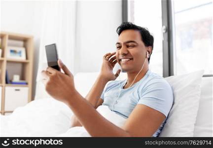 technology, bedtime and rest concept - happy smiling indian man in earphones with smartphone lying in bed and listening to music at home. indian man in earphones with phone in bed at home