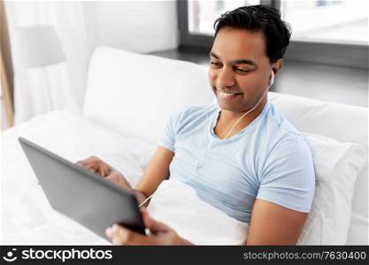 technology, bedtime and rest concept - happy smiling indian man in earphones with tablet pc computer lying in bed and listening to music at home. happy indian man with tablet pc in bed at home