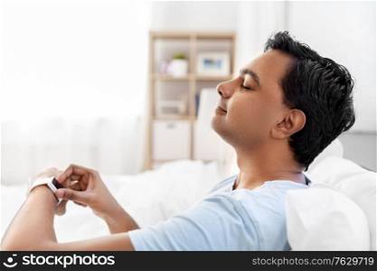 technology, bedtime and rest concept - happy smiling indian man with smart watch doing breath exercise lying in bed at home. happy indian man with smart watch in bed at home