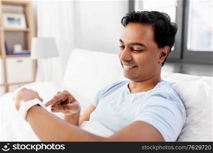 technology, bedtime and rest concept - happy smiling indian man with smart watch lying in bed at home. happy indian man with smart watch in bed at home