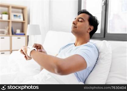 technology, bedtime and rest concept - happy smiling indian man with smart watch doing breath exercise lying in bed at home. happy indian man with smart watch in bed at home