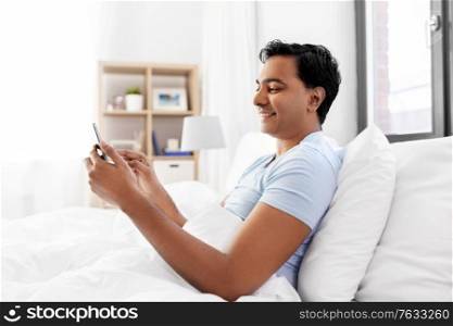 technology, bedtime and rest concept - happy smiling indian man with smartphone lying in bed at home. happy indian man with smartphone in bed at home