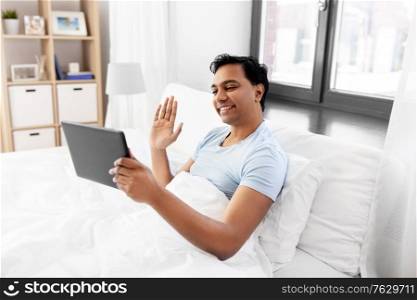 technology, bedtime and rest concept - happy smiling indian man with tablet pc computer lying in bed and having video call at home. happy man with tablet pc in bed having video call