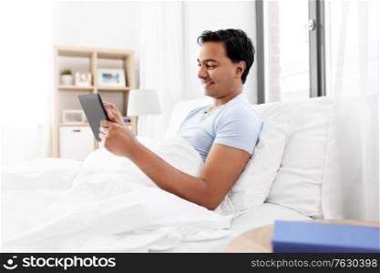 technology, bedtime and rest concept - happy smiling indian man with tablet pc computer lying in bed at home. happy indian man with tablet pc in bed at home