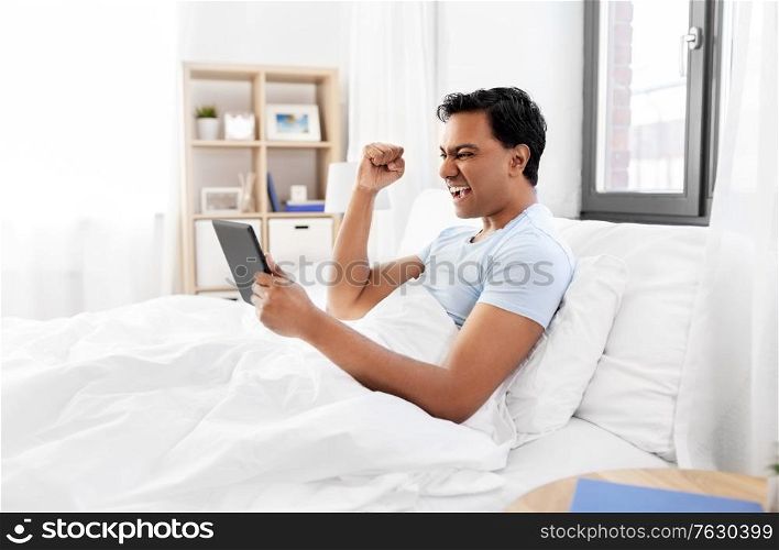 technology, bedtime and rest concept - happy smiling indian man with tablet pc computer lying in bed and making winning gesture at home. happy indian man with tablet pc in bed at home