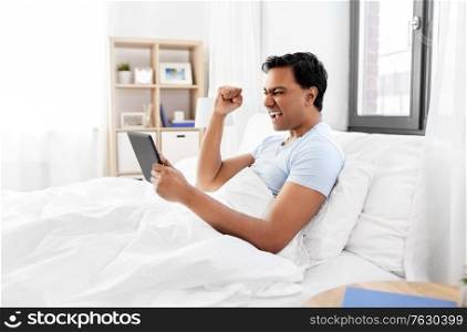 technology, bedtime and rest concept - happy smiling indian man with tablet pc computer lying in bed and making winning gesture at home. happy indian man with tablet pc in bed at home