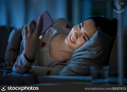 technology, bedtime and rest concept - happy smiling teenage girl in pajamas with smartphone lying in bed at night. teenage girl with smartphone lying in bed at night