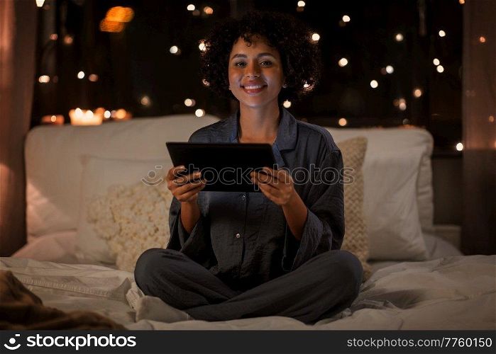 technology, bedtime and rest concept - happy smiling woman in pajamas with tablet pc computer sitting in bed at night. happy woman with tablet pc sitting in bed at night