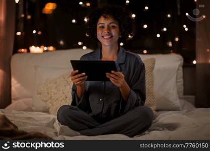 technology, bedtime and rest concept - happy smiling woman in pajamas with tablet pc computer sitting in bed at night. happy woman with tablet pc sitting in bed at night