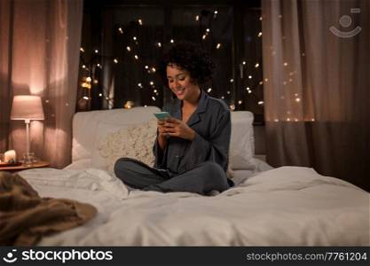technology, bedtime and rest concept - happy smiling woman in pajamas with smartphone sitting in bed at night. happy woman in pajamas with phone in bed at night