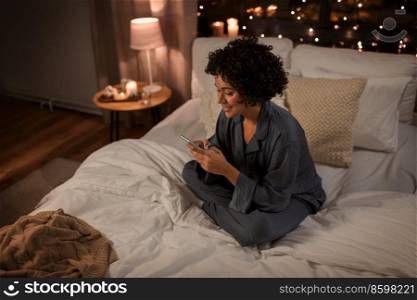 technology, bedtime and rest concept - happy smiling woman in pajamas with smartphone sitting in bed at night. happy woman in pajamas with phone in bed at night