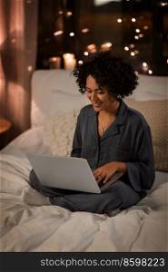 technology, bedtime and rest concept - happy smiling woman in pajamas with laptop computer sitting in bed at night. happy woman with laptop sitting in bed at night