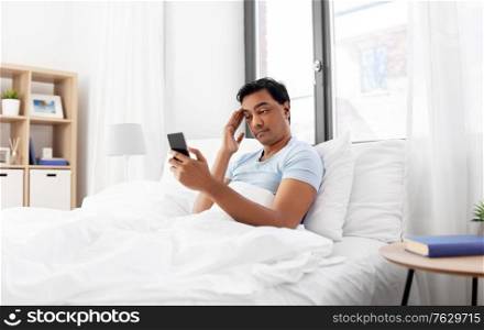 technology, bedtime and rest concept - sleepy indian man in bed looking at smartphone lying on table at home. sleepy indian man in bed looking at smartphone