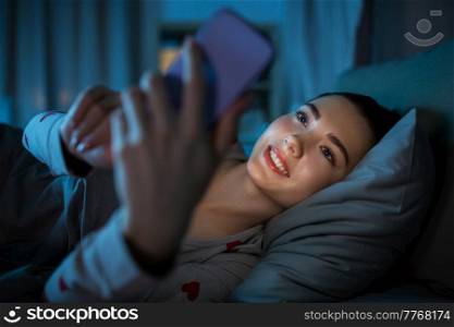 technology, bedtime and rest concept - smiling teenage girl in pajamas with smartphone lying in bed at night. teenage girl with phone lying in bed at night