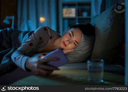 technology, bedtime and rest concept - teenage girl in pajamas with smartphone lying in bed at night. teenage girl with phone lying in bed at night