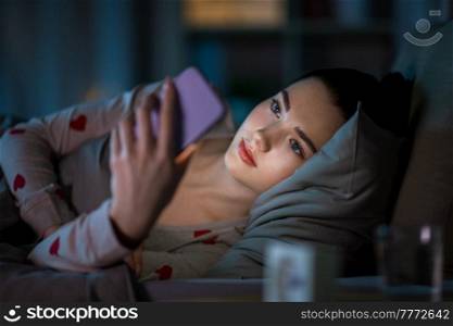technology, bedtime and rest concept - teenage girl in pajamas with smartphone lying in bed at night. teenage girl with smartphone lying in bed at night