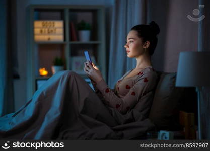 technology, bedtime and rest concept - teenage girl in pajamas with smartphone sitting in bed at night. teenage girl in pajamas with phone in bed at night