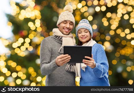 technology, christmas and winter clothes concept - happy couple in knitted hats and scarves with tablet computer over festive lights background. couple with tablet computer over christmas lights