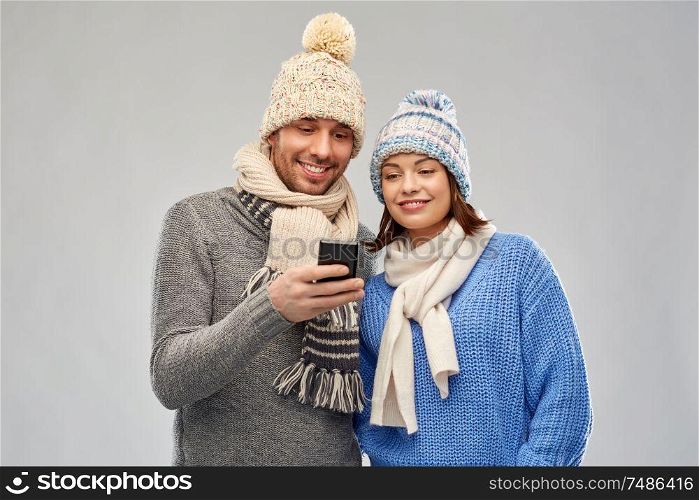 technology, christmas and winter clothes concept - happy couple in knitted hats and scarves with smartphone over grey background. happy couple in winter clothes with smartphone