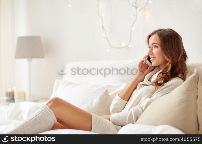 technology, christmas, communication and people concept - happy young woman lying in bed and calling on smartphone at home bedroom