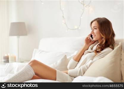 technology, christmas, communication and people concept - happy young woman lying in bed and calling on smartphone at home bedroom