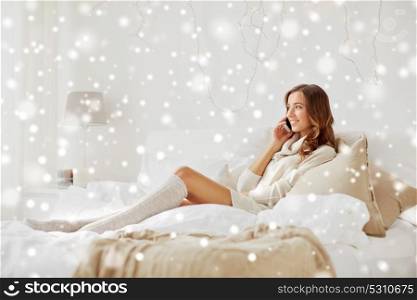 technology, christmas, communication and people concept - happy young woman lying in bed and calling on smartphone at home bedroom with snow. happy young woman with smartphone in bed at home