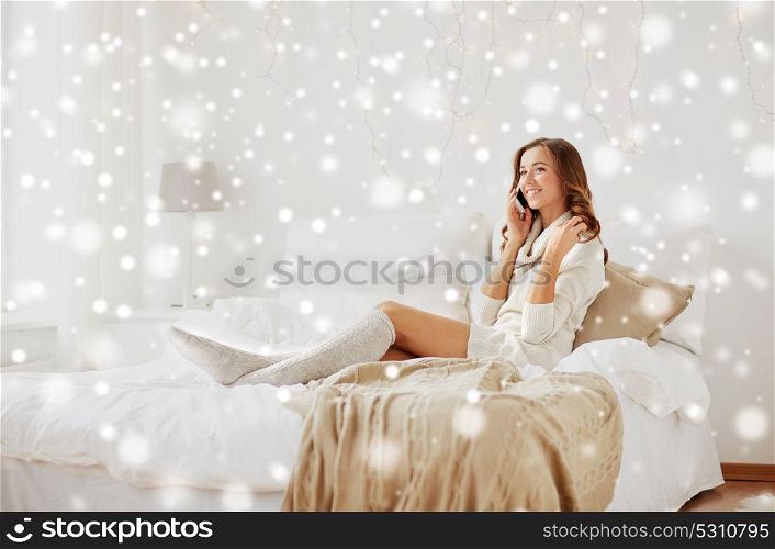 technology, christmas, communication and people concept - happy young woman lying in bed and calling on smartphone at home bedroom over snow. happy young woman with smartphone in bed at home