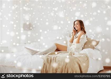 technology, christmas, communication and people concept - happy young woman lying in bed and calling on smartphone at home bedroom over snow. happy young woman with smartphone in bed at home