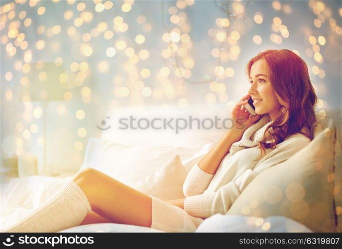 technology, christmas, communication and people concept - happy young woman lying in bed and calling on smartphone at home bedroom. happy young woman with smartphone in bed at home