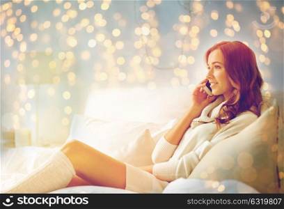 technology, christmas, communication and people concept - happy young woman lying in bed and calling on smartphone at home bedroom. happy young woman with smartphone in bed at home