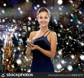 technology, christmas, holidays and people concept - smiling woman in evening dress holding smartphone over snowy night city background