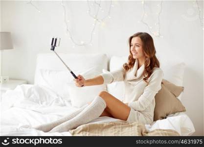 technology, christmas, winter and people concept - happy young woman in bed taking picture by smartphone selfie stick at home bedroom