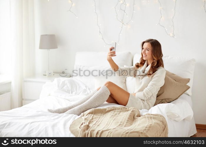 technology, christmas, winter and people concept - happy young woman in bed taking selfie on smartphone at home bedroom