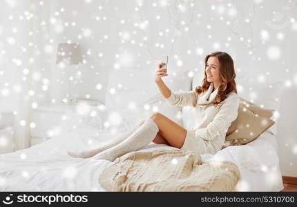 technology, christmas, winter and people concept - happy young woman in bed taking selfie on smartphone at home bedroom with snow. happy woman taking selfie by smartphone at home