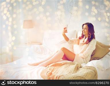 technology, christmas, winter and people concept - happy young woman in bed taking selfie on smartphone at home bedroom. happy woman taking selfie by smartphone at home