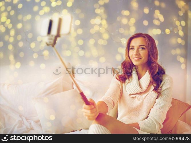 technology, christmas, winter and people concept - happy young woman in bed taking picture by smartphone selfie stick at home bedroom. happy woman taking selfie by smartphone at home