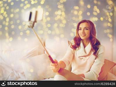 technology, christmas, winter and people concept - happy young woman in bed taking picture by smartphone selfie stick at home bedroom. happy woman taking selfie by smartphone at home