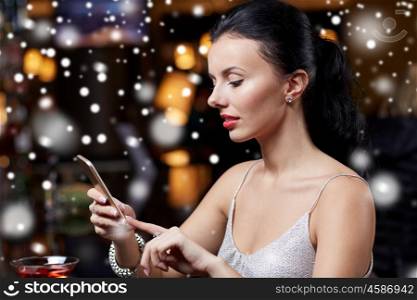 technology, christmas, winter holidays and people concept - young woman texting on smartphone at night club or bar over snow