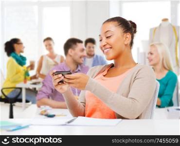 technology, clothing and tailoring concept - smiling african american female fashion designer with smartphone over studio and colleagues on background. fashion designer with smartphon over studio