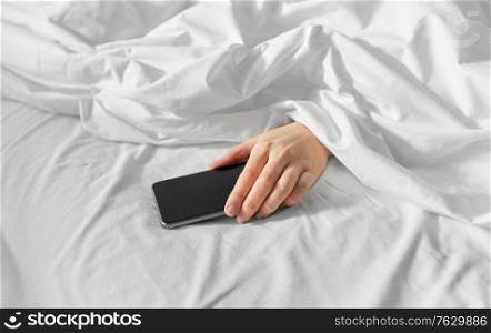 technology, comfort and morning concept - hand of young woman lying in bed with smartphone at bedroom. hand of woman lying in bed with smartphone