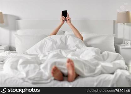 technology, comfort and morning concept - hands of young woman lying in bed with smartphone at bedroom. hands of woman lying in bed with smartphone