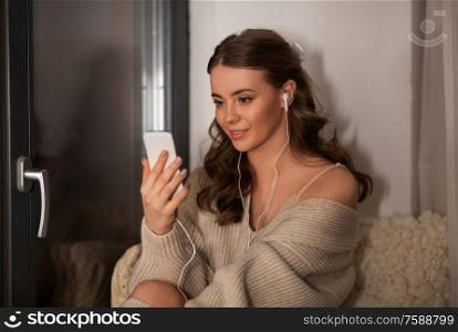technology, comfort and people concept - young woman in pullover with smartphone and earphones listening to music sitting at window at home. woman with smartphone and earphones at home