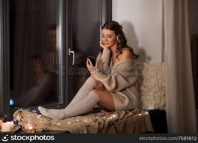 technology, comfort and people concept - young woman in pullover with smartphone and earphones listening to music sitting on windowsill at home. woman with smartphone and earphones at home