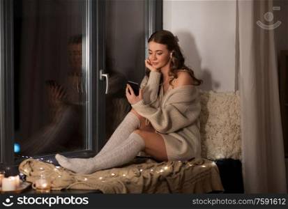 technology, comfort and people concept - young woman in pullover with smartphone and earphones listening to music sitting on windowsill at home. woman with smartphone and earphones at home