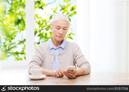 technology, communication age and people concept - senior woman with smartphone and coffee sitting at table and texting message at home over green natural background. senior woman with smartphone texting at home